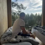 KoloCabins mökki sisältäpäin kuvattuna. Mies istuu sängyllä ja katselee maisemaa mökistä.